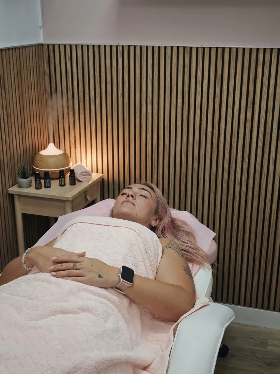 Clienta relajada en la camilla bajo una manta rosa, junto a un difusor de aromas y frascos de aceites esenciales